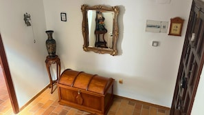 A room with a wooden chest, a wall-mounted mirror, a small table with a vase, and a wall-mounted clock.