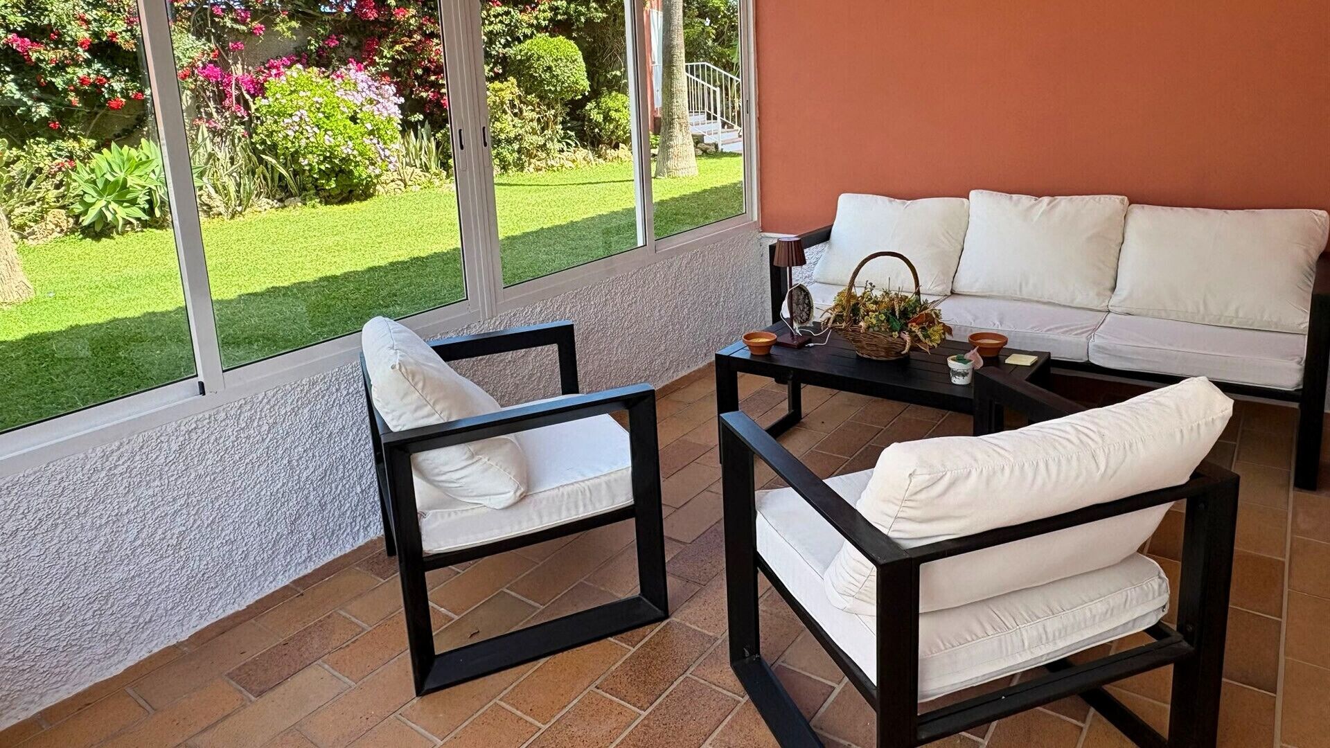 Une terrasse avec deux chaises à armature noire et un canapé, une petite table et une vue sur un jardin où poussent diverses plantes et fleurs.