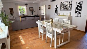 A dining area with a glass-top table, white chairs, and a grey sofa in the background.