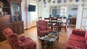 A living room with a wooden cabinet, a television, a dining area with chairs, and a kitchen in the background.