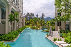 A swimming pool area with a curved design, surrounded by lush greenery and modern buildings. - The Title Legendary Designer 2BR near Bang Tao Beach (Choeng Thale)