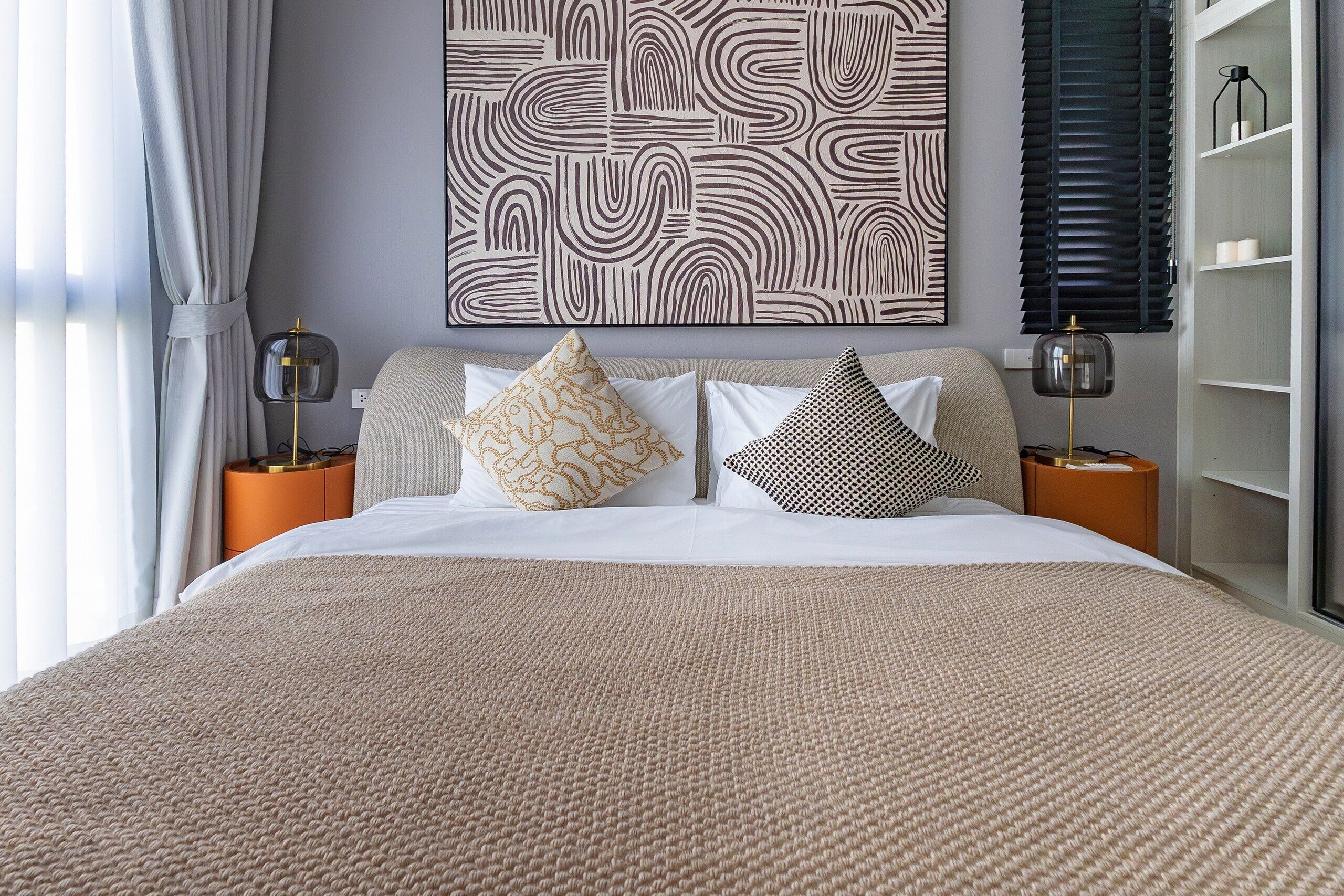 Una cama bien hecha con una manta texturizada, dos mesitas de noche con lámparas y una pared con un cuadro abstracto grande.