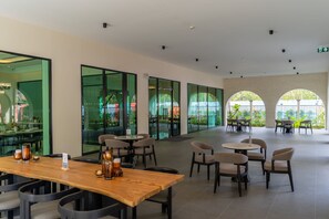 A spacious indoor area with multiple seating arrangements, including tables and chairs, and large windows offering views of greenery outside. - The Title Legendary Designer 2BR near Bang Tao Beach (Tambon Choeng Thale)