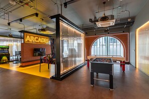 A modern indoor space with a foosball table, arcade gaming area, and a large window providing natural light. - The Title Legendary Designer 2BR near Bang Tao Beach (Tambon Choeng Thale)