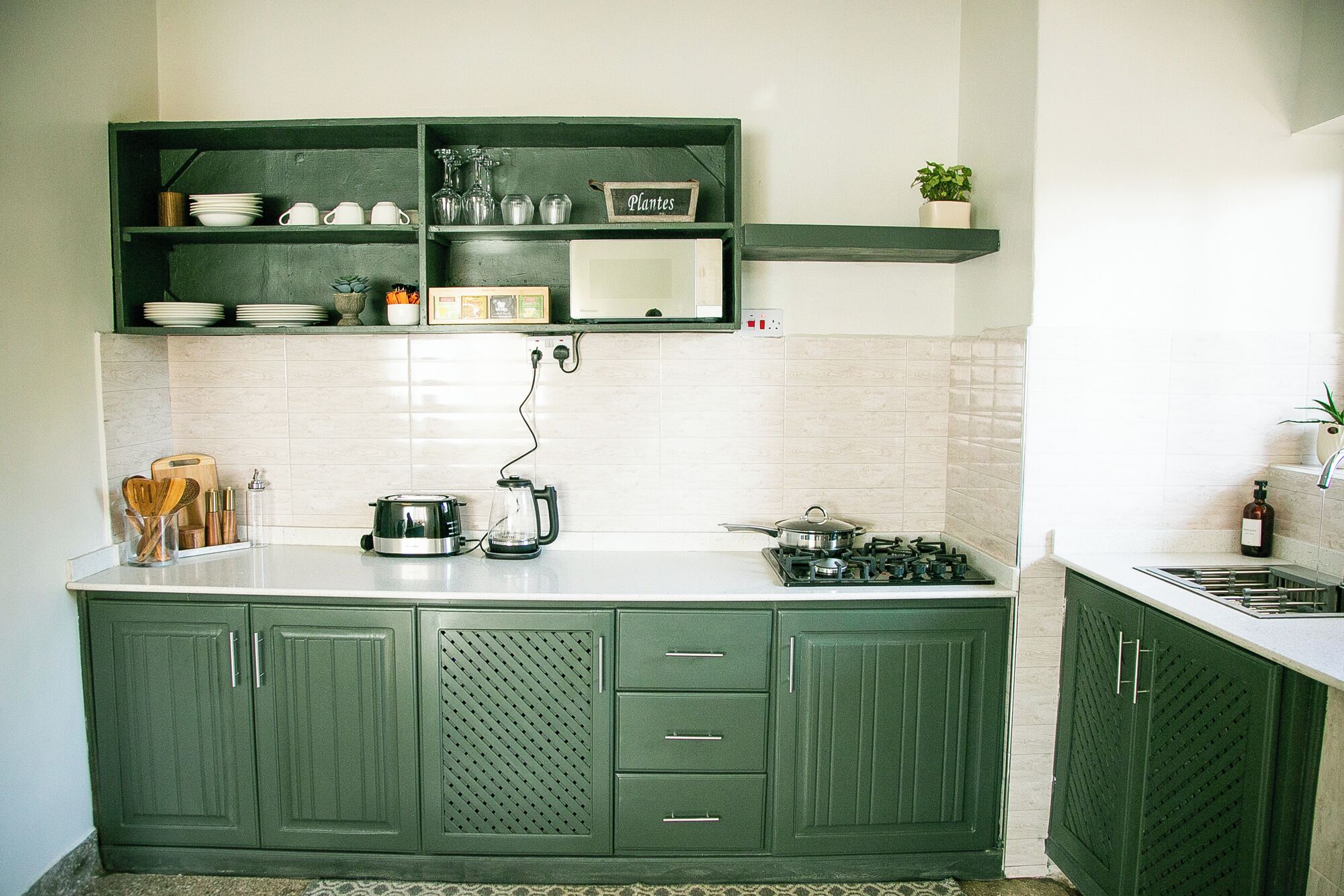 A kitchen with green cabinets, a white countertop, and a small shelf with glassware and a sign labeled 'Plantes'.