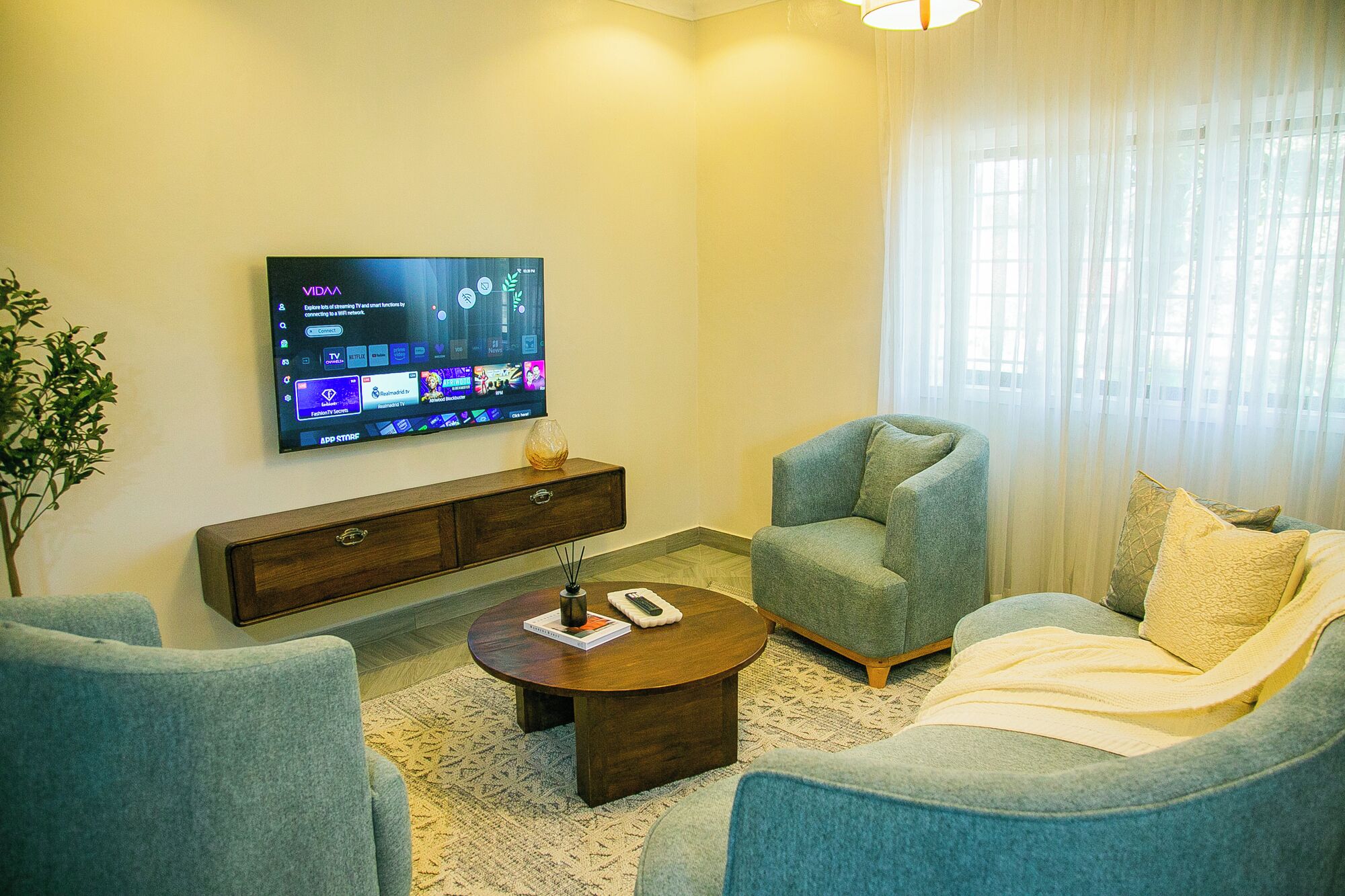 A living room with a flat-screen TV mounted on the wall, a wooden console table, a round coffee table, two armchairs, and a sofa.
