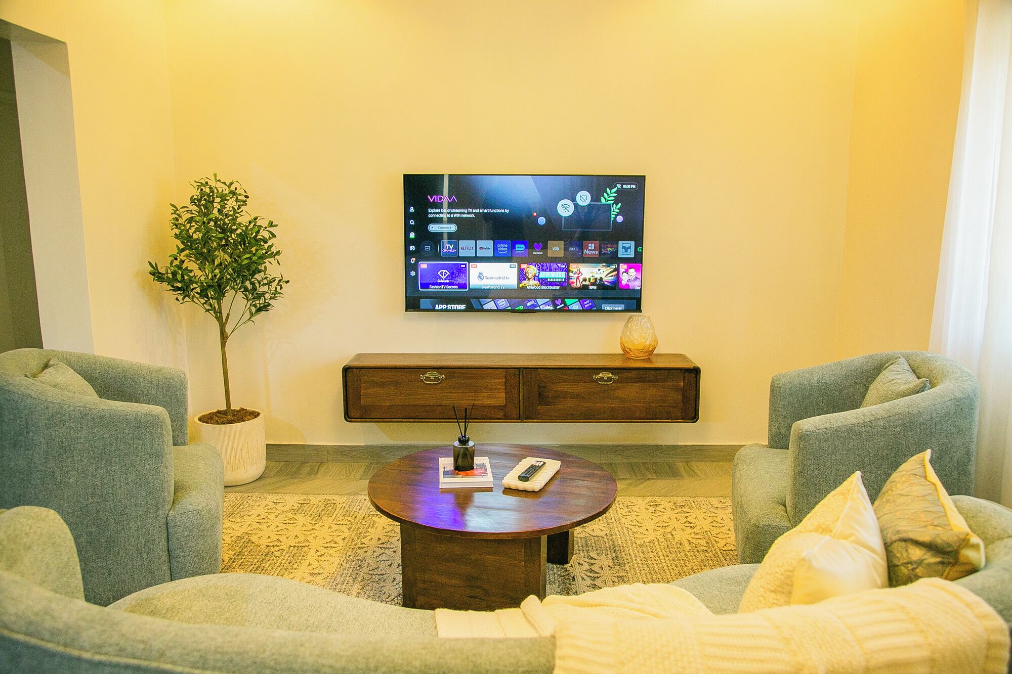 Een woonkamer met een flatscreen televisie aan de muur, een houten kast eronder, twee fauteuils, een salontafel met een afstandsbediening en een boek, en een potplant.