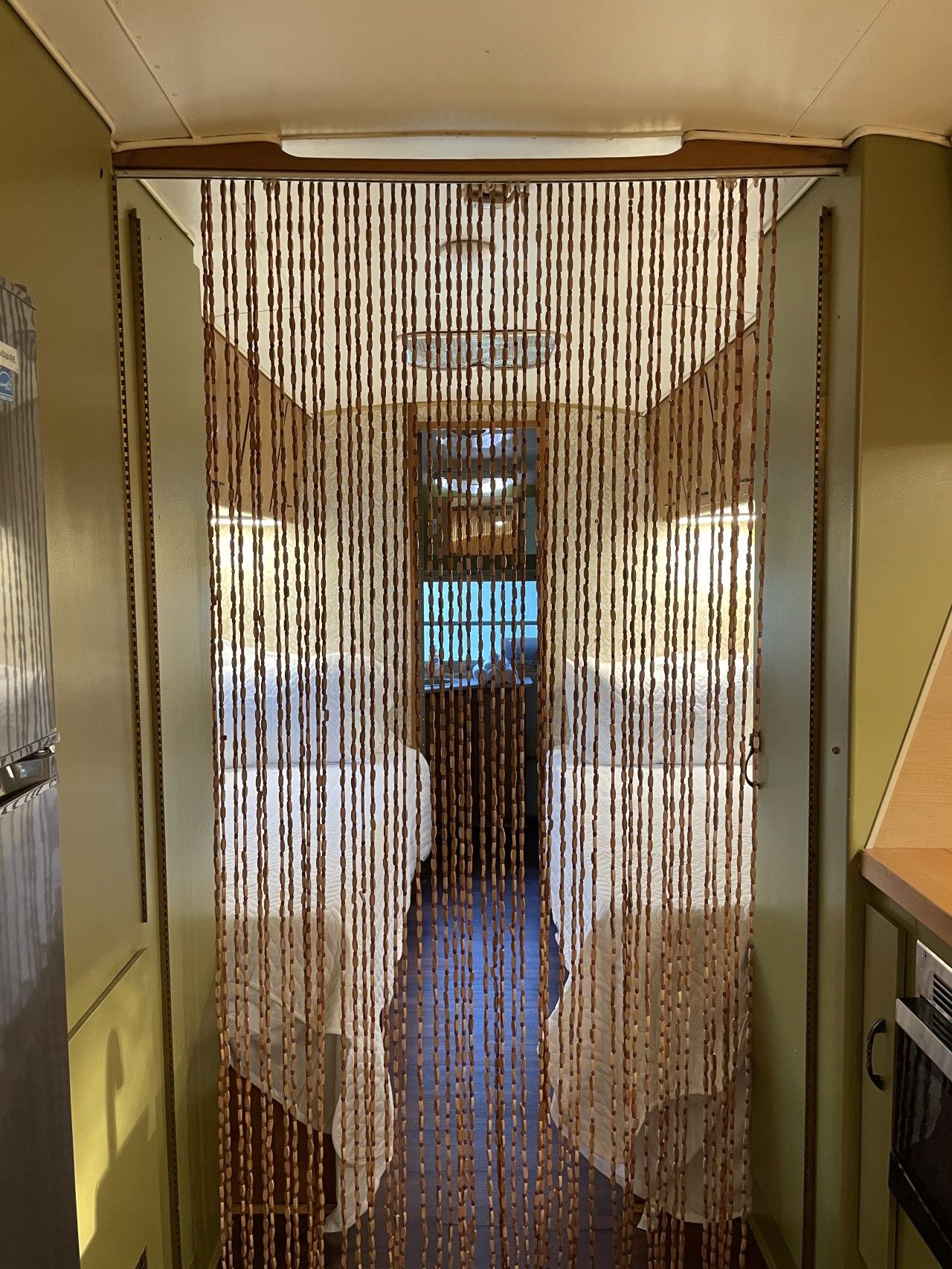 Un pasillo estrecho con una serie de persianas verticales, una kitchenette a la derecha y una cama con cabecera visible entre las persianas.