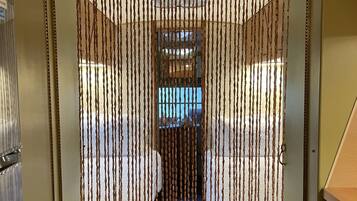 Un pasillo estrecho con una serie de persianas verticales, una kitchenette a la derecha y una cama con cabecera visible entre las persianas.