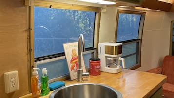 Une cuisine compacte dotée d’un évier en acier inoxydable, d’une machine à café et d’une fenêtre donnant sur les arbres.