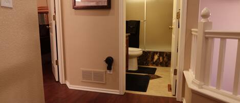 A hallway leading to a bathroom with a toilet, a shower, and a vent on the wall.