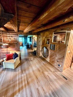 A log cabin interior with a wooden floor, a sofa with cushions, a round mirror, and a window with a view.