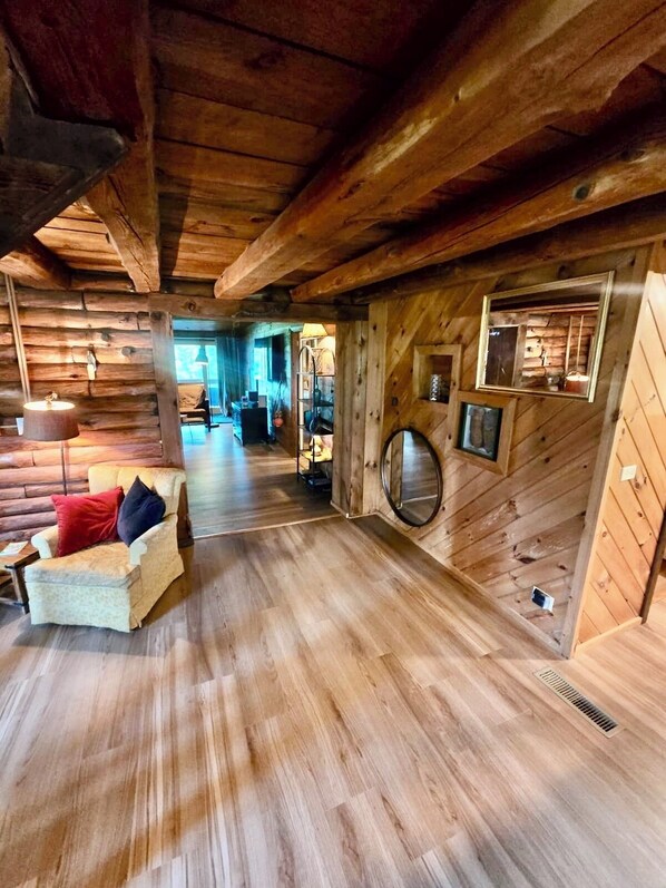 A log cabin interior with a wooden floor, a sofa with cushions, a round mirror, and a window with a view.