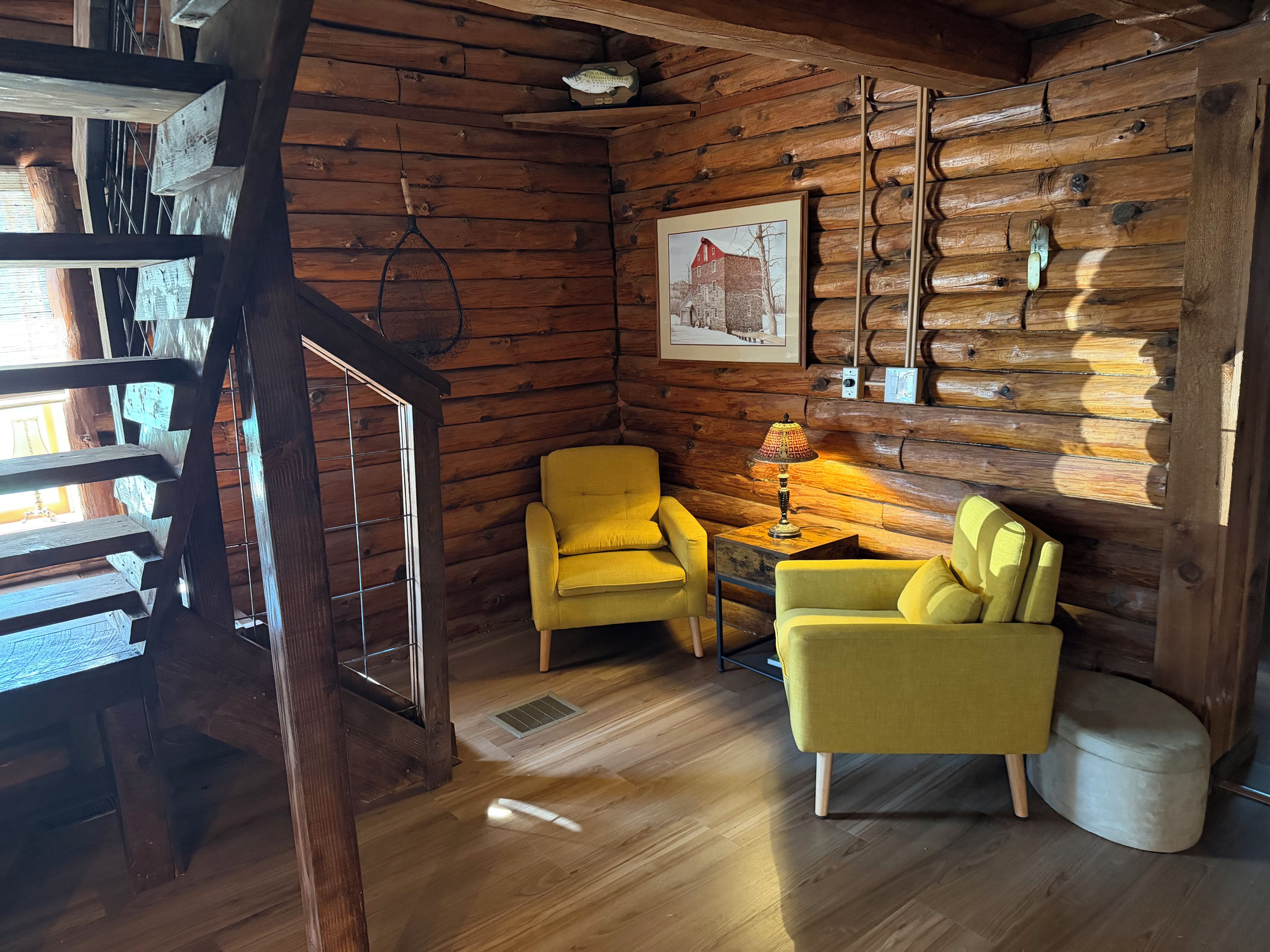 L'intérieur d'une cabane en rondins, avec un escalier en bois, deux fauteuils jaunes, une table avec une lampe et un tableau encadré accroché au mur.