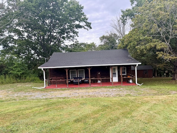 A log cabin with a porch, a covered picnic table, and a small shed in a grassy area.