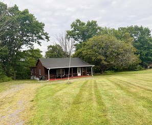 A rustic log cabin with a porch, surrounded by a well-maintained lawn and dense trees.