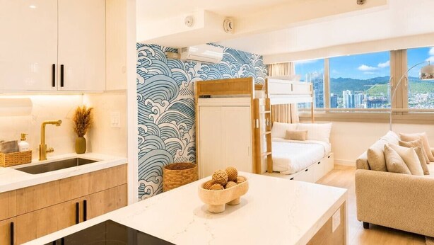 A modern kitchen with a central island, a dining area with a sofa, and a view of a cityscape through large windows. - Luxe Island Retreat – Modern Coastal Design (Honolulu)