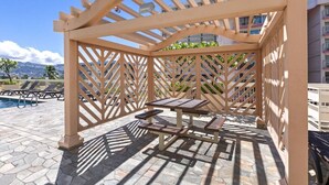 A wooden pergola structure with a table and benches, overlooking a swimming pool area. - Luxe Island Retreat – Modern Coastal Design (Honolulu)