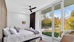 Ein modernes Schlafzimmer mit Bett, Deckenventilator, großem Fenster und Blick ins Freie.