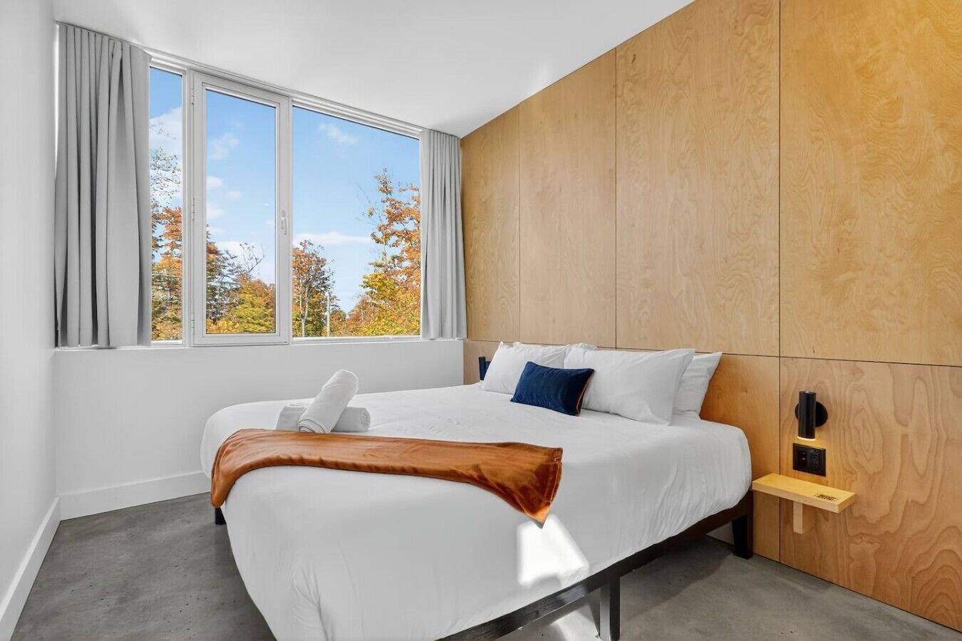 Une chambre d’hôtel avec un grand lit, une tête de lit en bois, une fenêtre avec des rideaux et une table de chevet avec une lampe.
