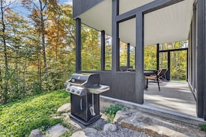 A modern outdoor patio with a built-in grill, surrounded by a wooden deck and a view of autumn foliage. - Ultra Luxury Chalet: Private Oasis, Spa, Sauna (Les Laurentides Regional County Municipality)