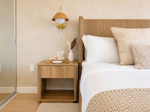 A bedroom with a wooden nightstand, a bedside lamp, a bed with white bedding, and a textured rug. - Waikiki Ocean Breeze Escape: Coastal Luxe (Honolulu)