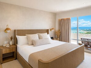 A modern hotel bedroom with a large bed, wooden headboard, bedside lamps, a nightstand, and a view of the city through large windows. - Waikiki Ocean Breeze Escape: Coastal Luxe (Honolulu)
