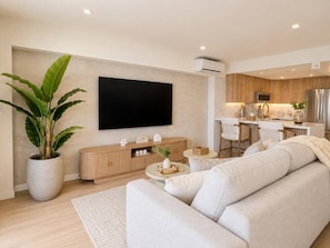 Un salon moderne avec un téléviseur à écran plat, une grande plante en pot, un canapé et un coin cuisine à l’arrière-plan.