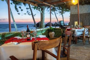 Un comedor con una mesa dispuesta para dos personas, con vista al océano, palmeras y un atardecer.