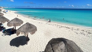 En strand med hvidt sand, krystalklart turkis vand og stråtagsparasoller.
