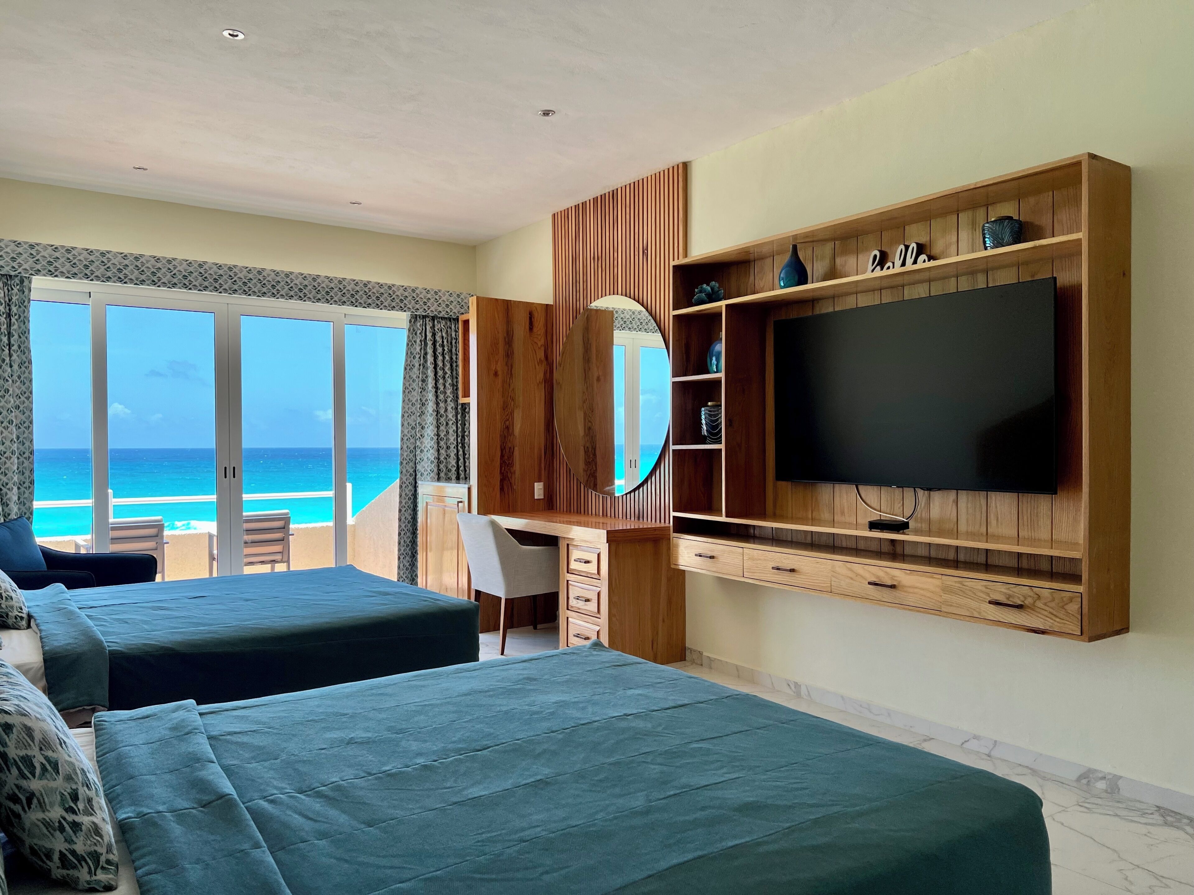 A hotel room with two beds, a TV, a desk, and a large window overlooking the ocean.