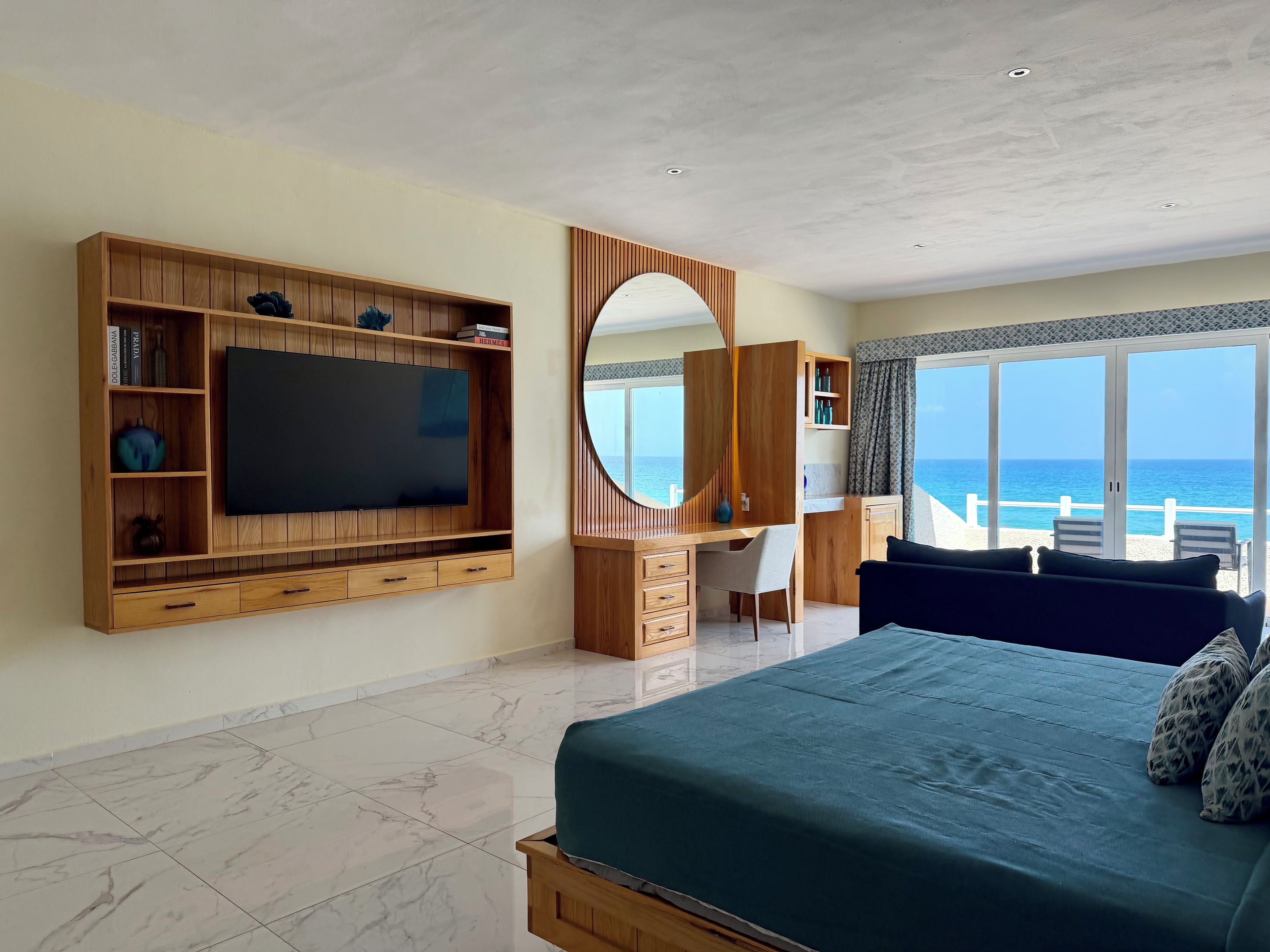 A bedroom with a large bed, a TV, a desk, and a view of the ocean.