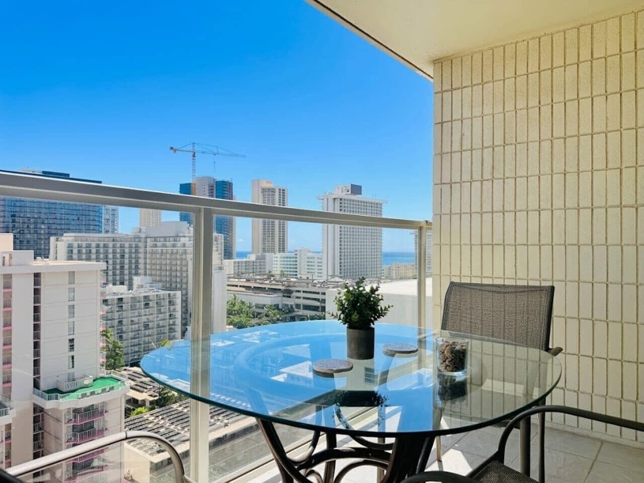 A balcony with a glass table and chairs, a potted plant, and a view of a cityscape.