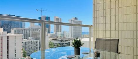 A balcony with a glass table and chairs, a potted plant, and a view of a cityscape.