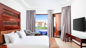 A hotel room with a bed, a desk, a chair, a television, and a view of a pool and buildings. - Pickalbatros Portofino Villaggio Resort (Marsa Alam)