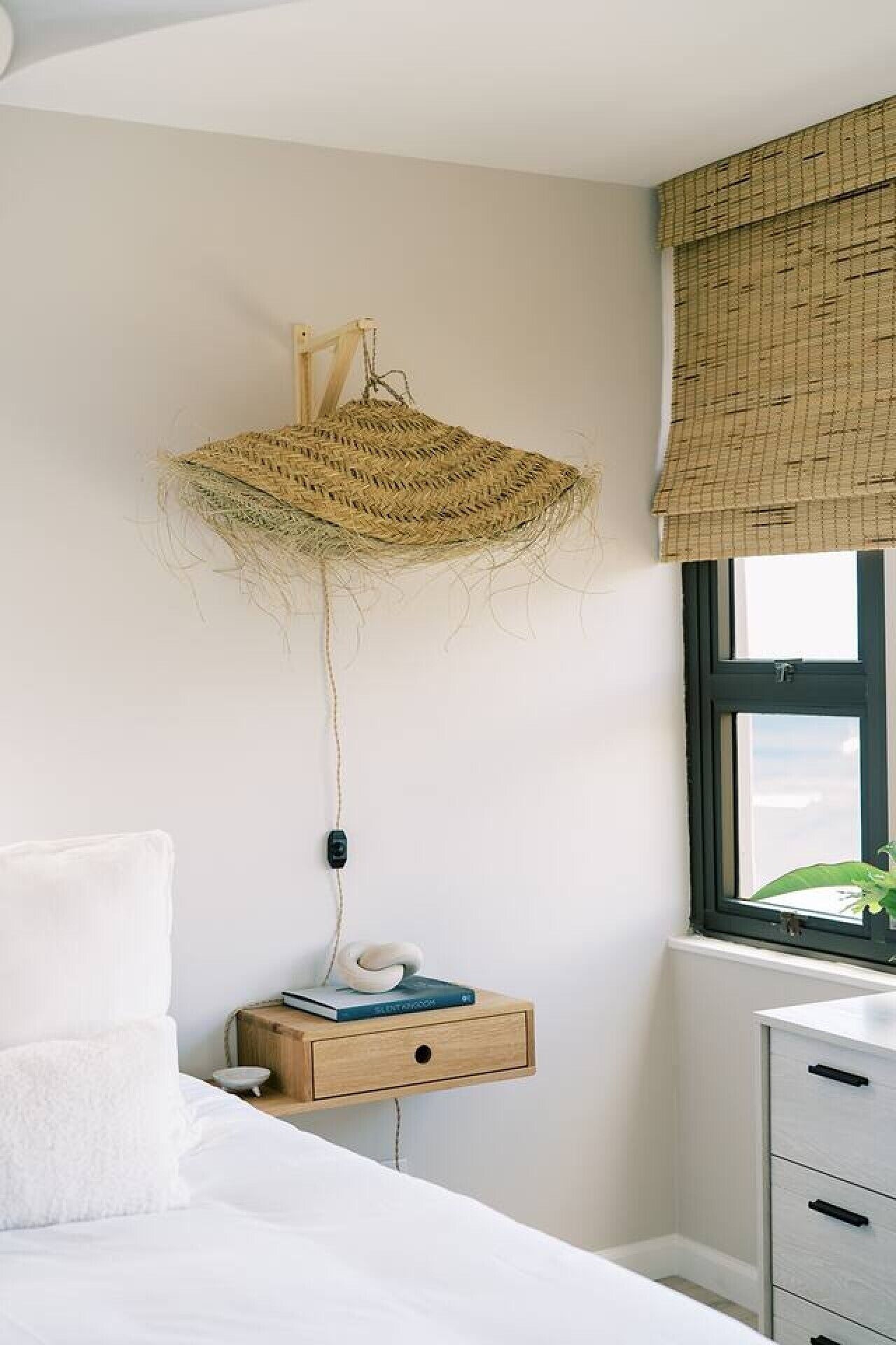 Ein Schlafzimmer mit einem Bett, einem Nachttisch mit einem Buch, einer wandmontierten gewebten Leuchte und einem Fenster mit Blick.