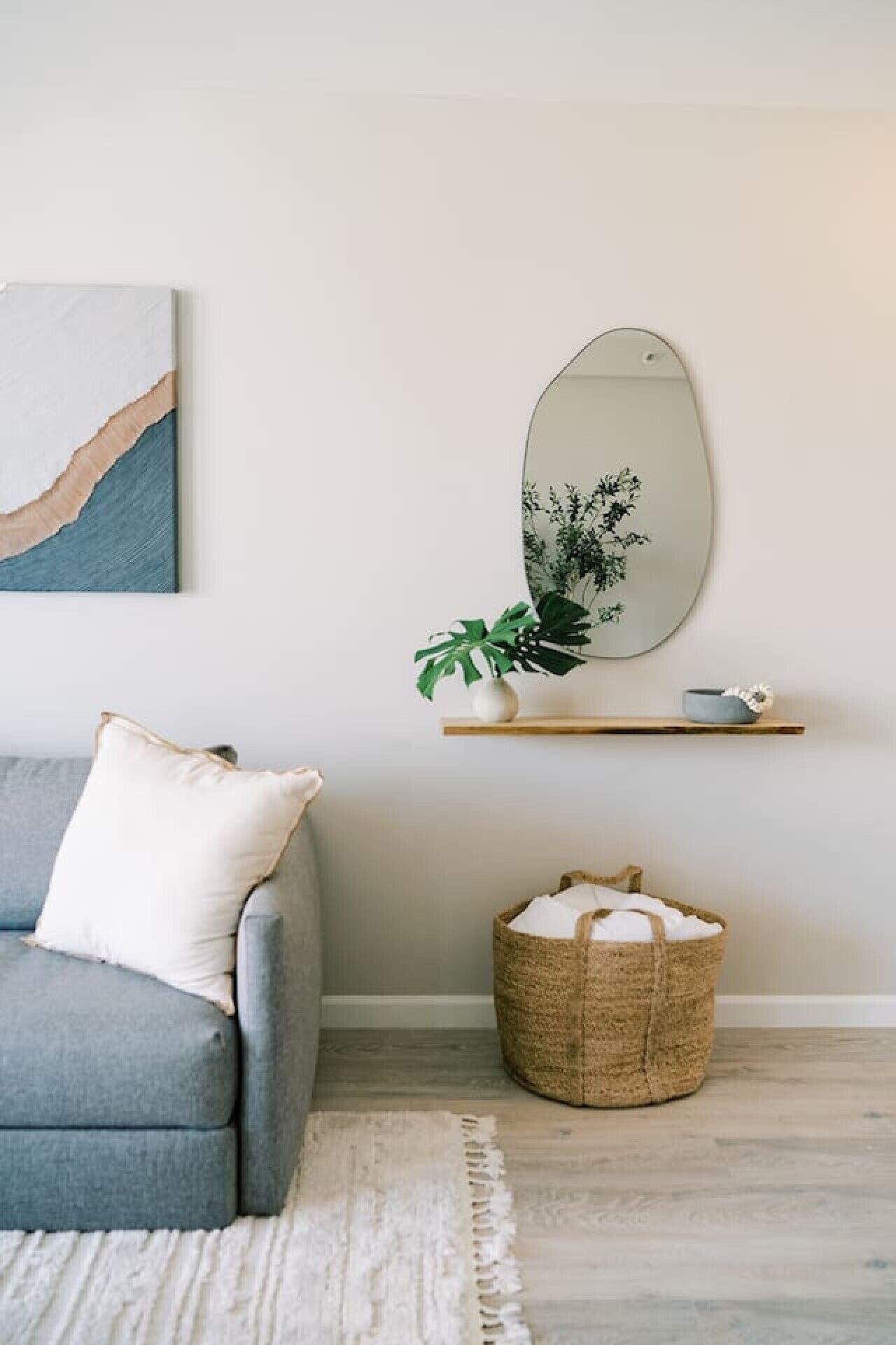 A living room with a grey sofa, a white cushion, a woven basket, a mirror, and a small shelf with a plant and a bowl.