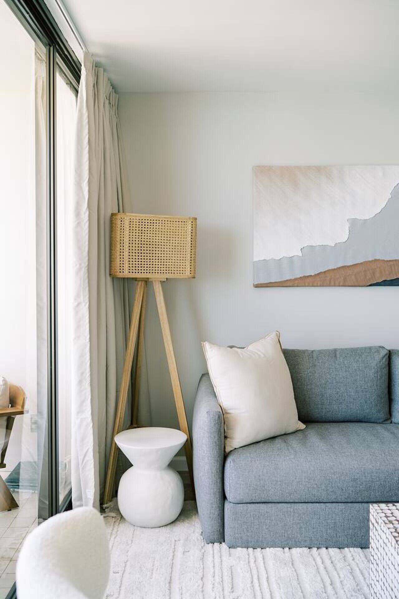 Ein modernes Wohnzimmer mit einem grauen Sofa, weißen Kissen, einem weißen runden Beistelltisch, einer gewobenen Stehlampe und einem großen abstrakten Gemälde an der Wand.