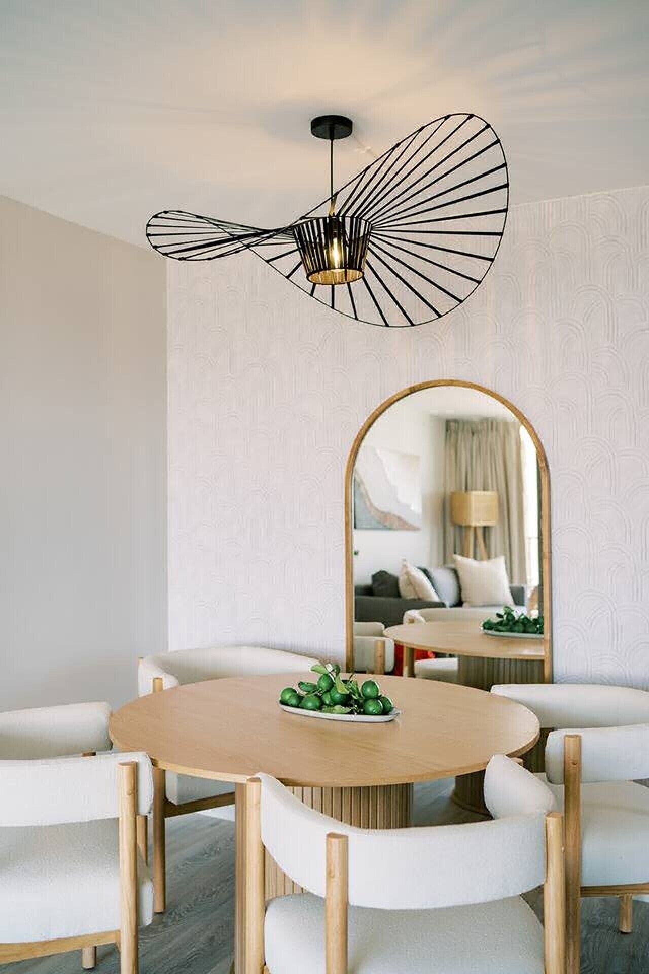 A dining area with a round table, white upholstered chairs, a modern pendant light, and a large mirror reflecting a room with a bed and a lamp.