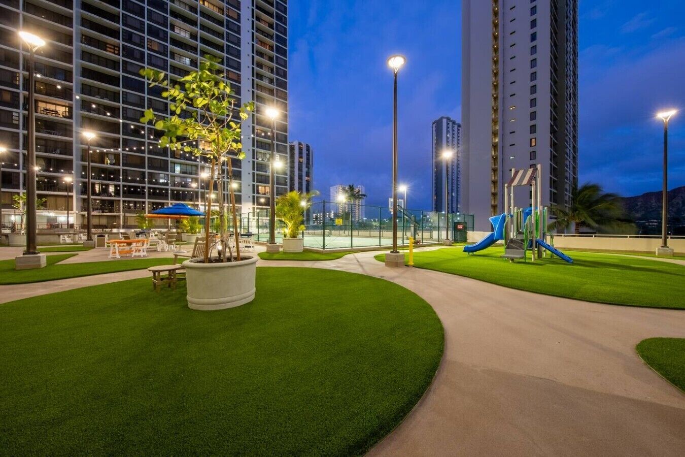 A well-lit outdoor area with artificial grass, a playground, and tall buildings in the background.