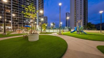 A well-lit outdoor area with artificial grass, a playground, and tall buildings in the background.