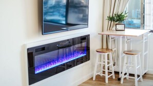 En moderne stue med et fladskærms-tv på væggen, en indbygget pejs med blå flammer, og to hvide barstole.
