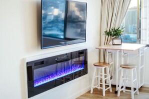 Ein modernes Wohnzimmer mit einem flachbildfernseher an der Wand, einem eingebauten Kamin mit blauem Feuer und zwei weißen Barhocker.
