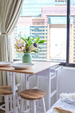 A modern room with a view of tall buildings, a table with two plates and a vase of flowers, and a window with curtains. - Modern Waikiki Studio Oasis (Honolulu)