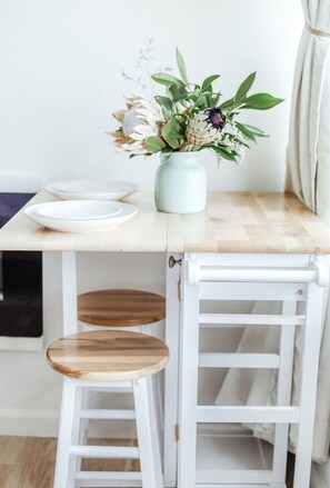 A small dining area with a white table, a vase with flowers, and two wooden stools. - Modern Waikiki Studio Oasis (Honolulu)