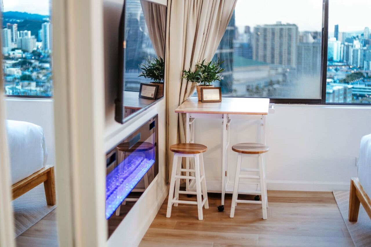 Ein modernes Zimmer mit Stadtblick, ein kleiner Tisch mit zwei Hocker, ein Fernseher und ein Regal mit Dekoration.