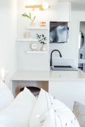 A modern kitchen with white cabinetry, a built-in microwave, a sink, and a small shelf with decorative items. - Modern Waikiki Studio Oasis (Honolulu)