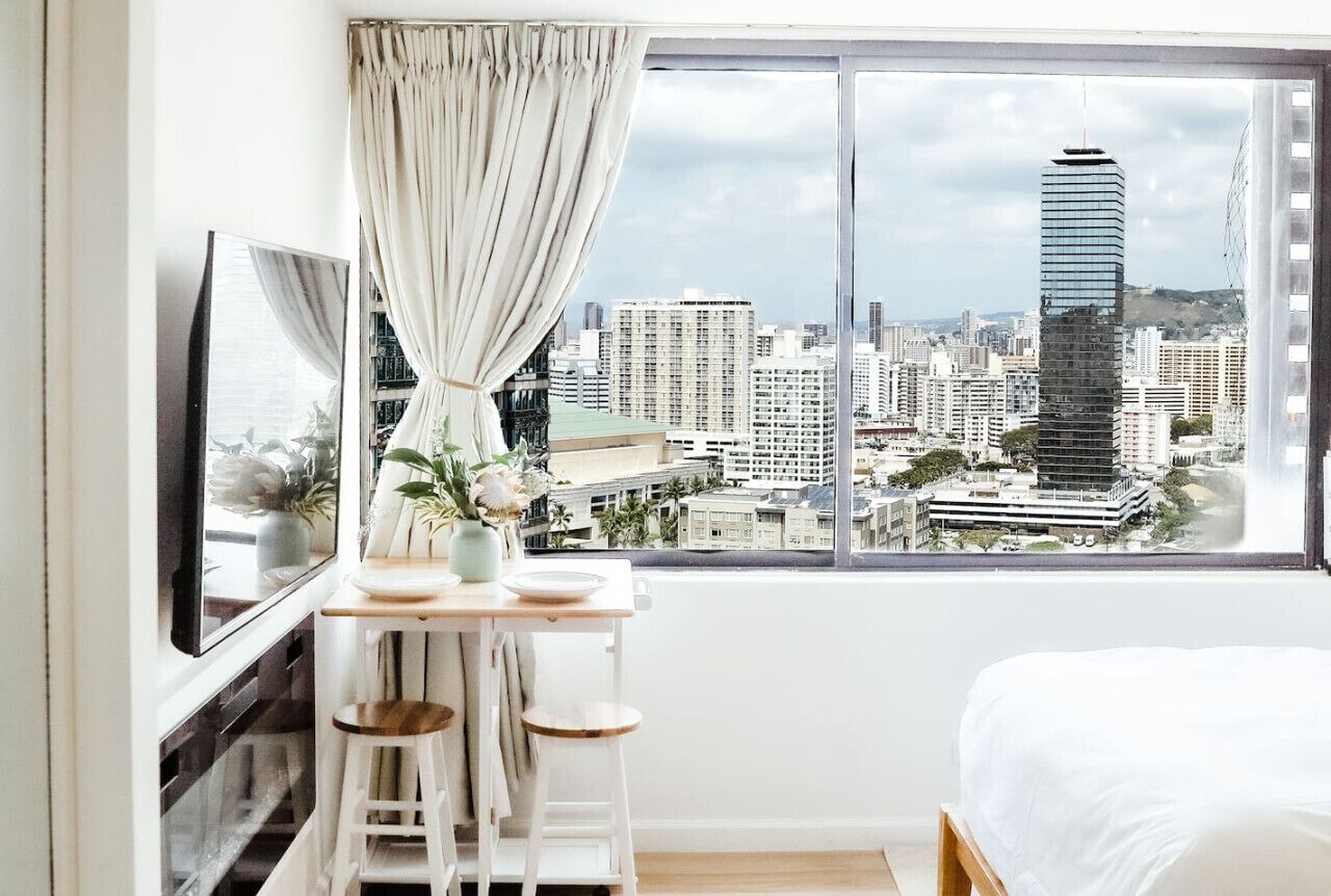 A bedroom with a bed, a TV, a table with chairs, a window with curtains, and a city view.