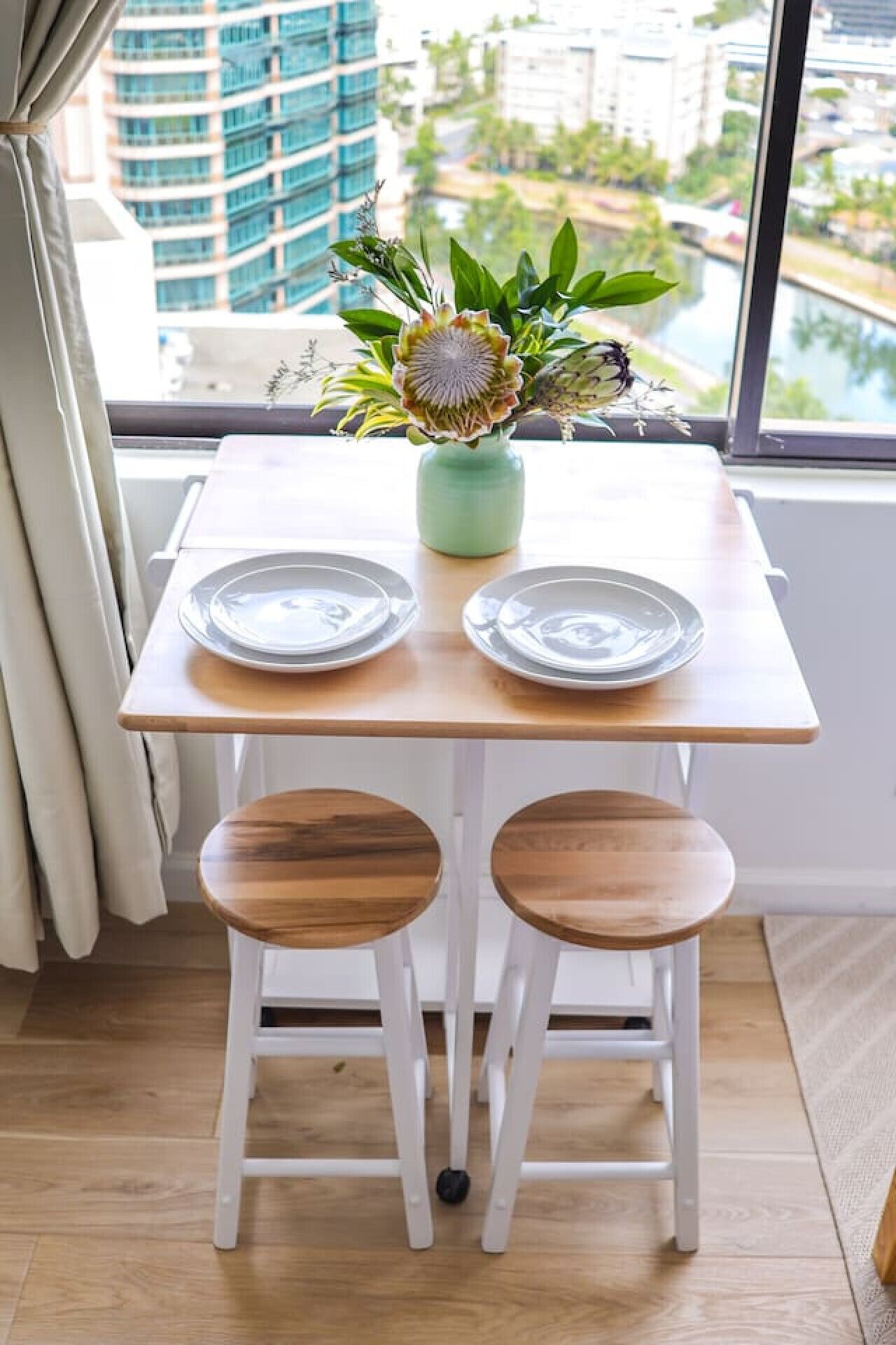 Een eettafel met twee stoelen, een vaas met een bloem, en uitzicht op gebouwen door een raam.