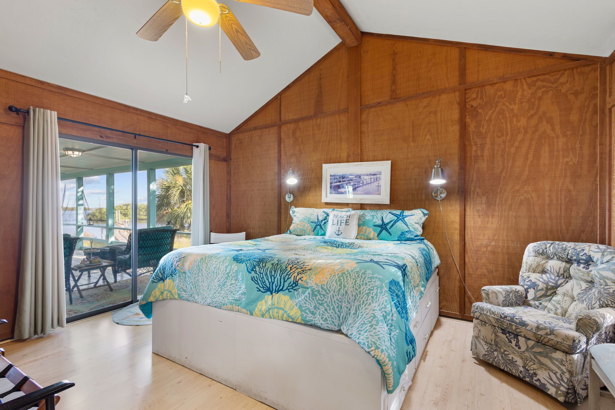 Quarto com cama, uma cadeira, ventilador de teto e vista através de uma porta de vidro.